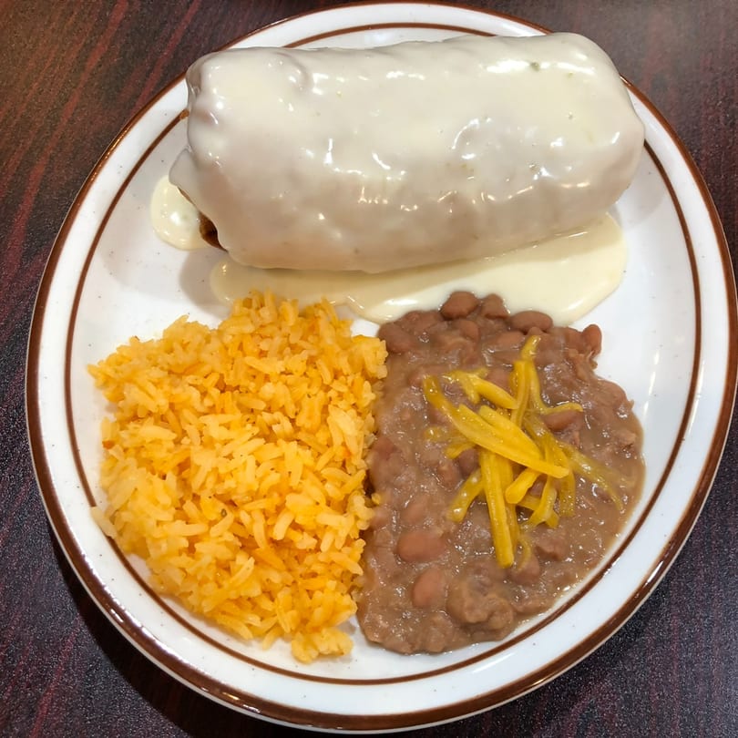 A breakfast burrito covered in white gravy, with a side of yellow rice and refried beans.