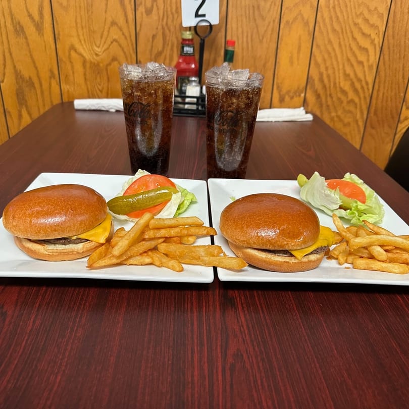 Two cheeseburger platters with fries, lettuce, tomato, pickle, and two Coca-Colas on a wooden table.